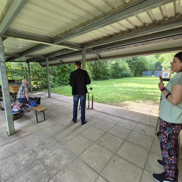 Drei Personen stehen auf einer überdachten Terrasse mit einem Tisch. Eine Person mit Pfeil und Bogen zielt auf eine Zielscheibe im Freien, umgeben von Bäumen.