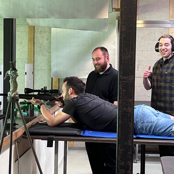 Ein Mann liegt auf dem Bauch und zielt mit einem Gewehr, während zwei andere Männer daneben stehen und zuschauen. Sie befinden sich in einem Schießstand.