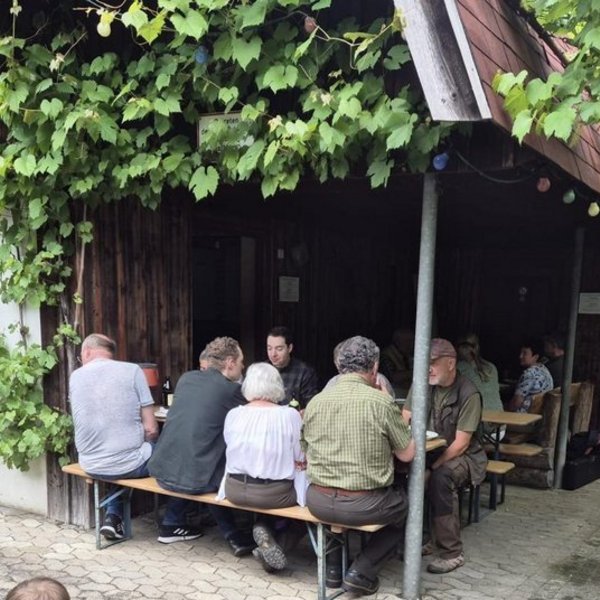 Personen sitzen an einem Holztisch vor einem mit Efeu bewachsenen Gebäude. Ein Kind fährt Fahrrad, während ein anderes mit Roller steht. Ein Hund liegt entspannt auf dem Boden.
