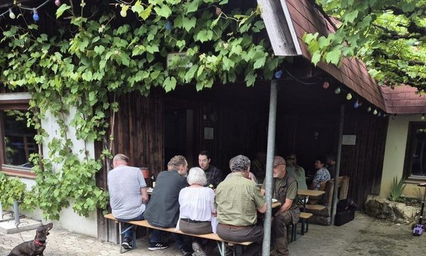 Personen sitzen an einem Holztisch vor einem mit Efeu bewachsenen Gebäude. Ein Kind fährt Fahrrad, während ein anderes mit Roller steht. Ein Hund liegt entspannt auf dem Boden.