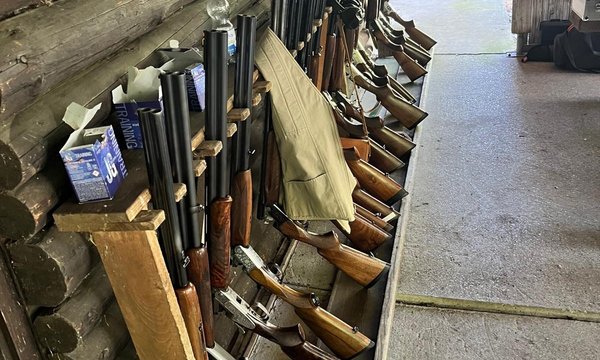 Gewehre stehen ordentlich in einem Holzständer in einer Hütte. Auf dem Boden liegt ein Teppich, helle Wände umgeben die Szene. Ein offener Eingang führt nach draußen. Text: „TRUST“ auf der Tür.